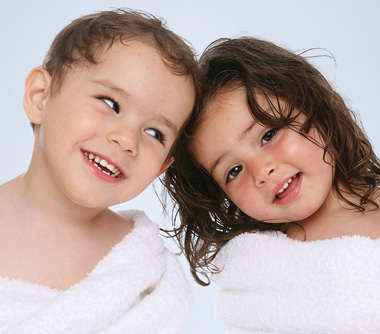 Image of a little boy and a little girl wrapped up on a white towel. Girl is looking ahead whilst oy is smiling and looking at the girl.